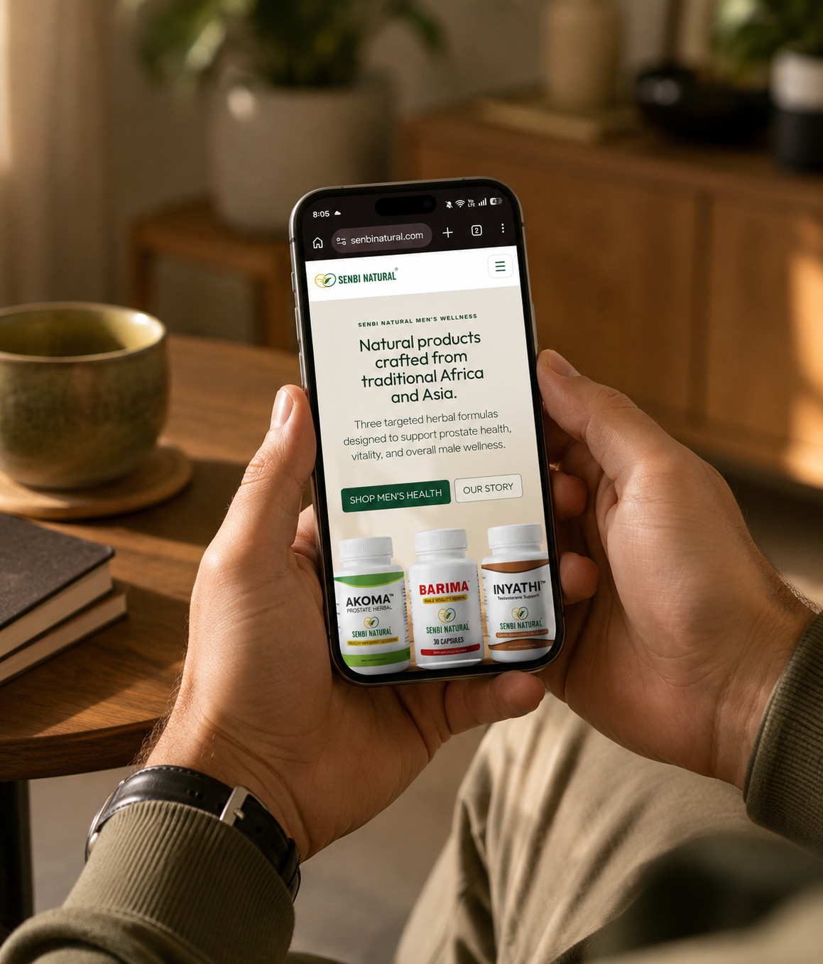 Masculine hands holding a smartphone displaying the Senbi Natural mobile homepage in a warm residential setting